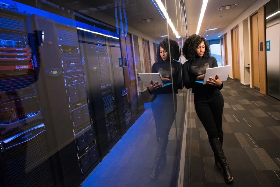 A practitioner walking a data centre aisle with a laptop, server racks lit blue beside her.